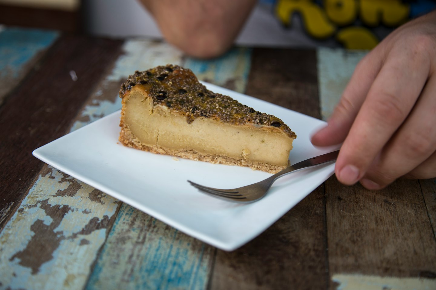 A person reaches for a fork beside a slice of cheesecake.